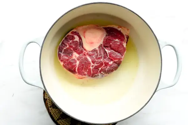 A thick beef shank, or osso buco in Spanish, is browning in a light-colored pot with oil.