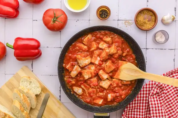 Tuna in tomato sauce simmering in a pan, with fresh tomatoes, red bell peppers, and crusty bread on the side.