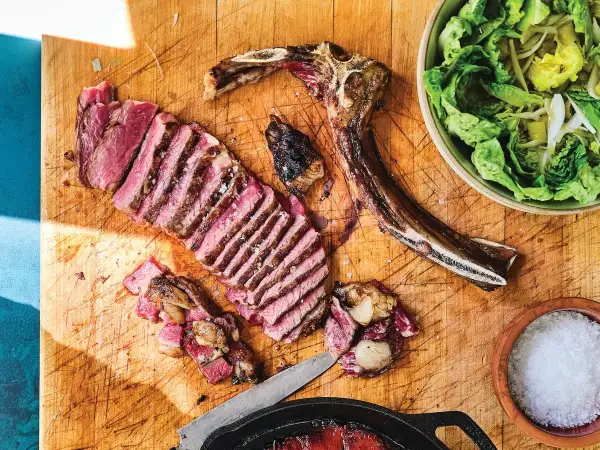 A perfectly cooked Spanish steak, sliced and ready to enjoy, sits on a wooden board with a side of fresh salad and a bowl of coarse salt.