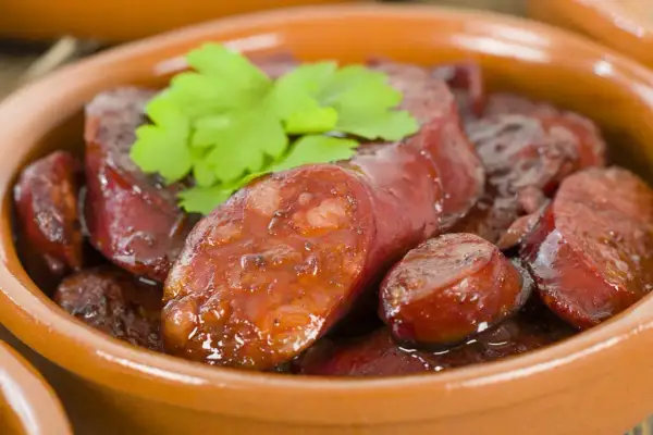 Slices of glistening chorizo al vino, simmered in a rich sauce, are served in a rustic clay bowl, garnished with fresh parsley.