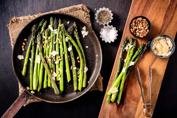A pan of cooked asparagus with shaved parmesan and pine nuts, showing how to make asparagus a delicious side dish.