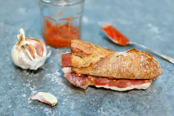 A delicious bocadillo recipe featuring a crusty baguette filled with cured ham and a vibrant tomato spread, with garlic and a jar of spread nearby.